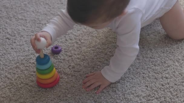Uma criança de um ano brincando com brinquedos educativos em casa. Menino com olhos azuis está brincando com o brinquedo da pirâmide no chão. Pequena criança se divertir dentro de casa — Vídeo de Stock