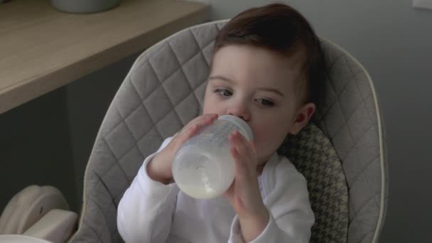 En pojke på ett år dricker mjölk ur sin flaska, sitter på en barnstol, livsstil familj morgon frukost — Stockvideo
