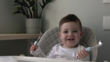 Bebeğim gülümsüyor, çocuk koltuğunda oturuyor ve yemeğe hazır, kaşık ve halk ile oynuyor, evde yaşam tarzı aile sabahı