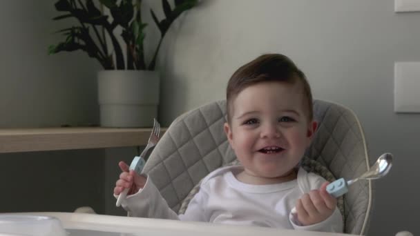 Baby pojke ler, sitter i en barnstol och redo att äta, leka med sked och folk, livsstil familj morgon hemma — Stockvideo