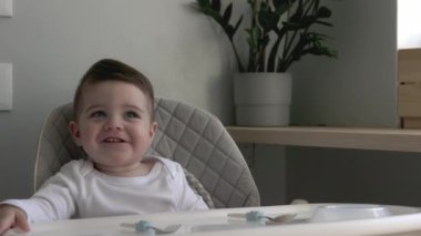 Bebeğim gülümsüyor, çocuk koltuğunda oturuyor ve yemeğe hazır, kaşık ve halk ile oynuyor, evde yaşam tarzı aile sabahı