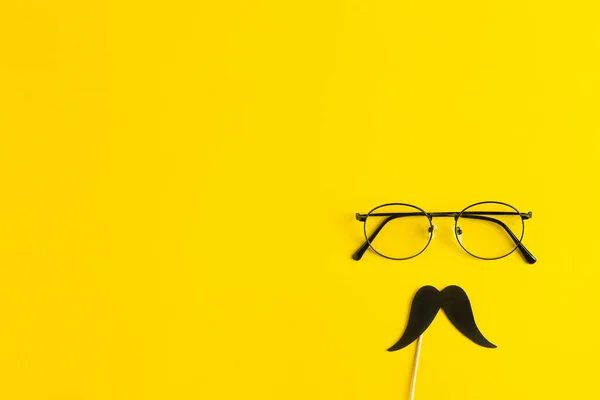 Ημέρα των πατέρων έννοια διακοπών. Διάφανα γυαλιά, κομψό μαύρο χάρτινο φωτογραφικό περίπτερο στηρίζει μουστάκια σε κίτρινο φόντο. — Φωτογραφία Αρχείου