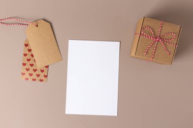 Sevgililer günü, hediyeden yapılmış anneler günü kompozisyonu, kırmızı çiçek, kalpli etiket kağıdı, bej arka planda beyaz kart. Düz konum, üst görünüm, kopyalama alanı.