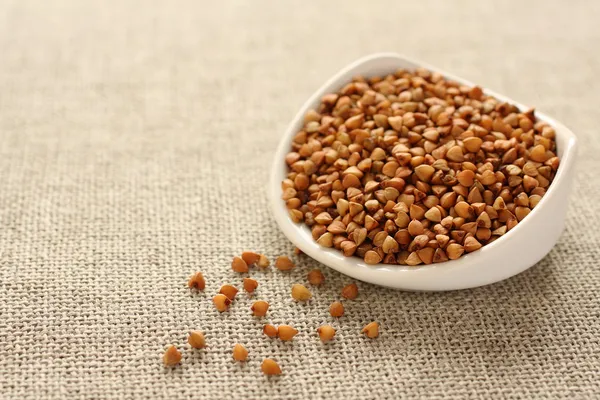 Buckwheat grains in white ceramic bowl on sackcloth background — Stock Photo, Image