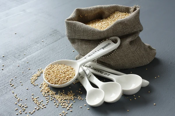Quinoa grain in small burlap sack and porcelain measuring spoons — Stock Photo, Image