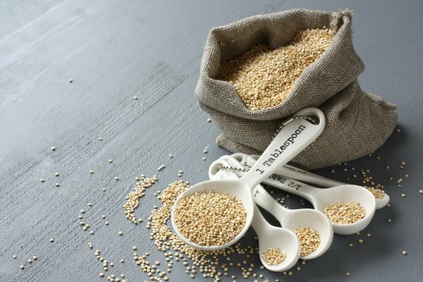Quinoa grain in small burlap sack and porcelain measuring spoons — Stock Photo, Image