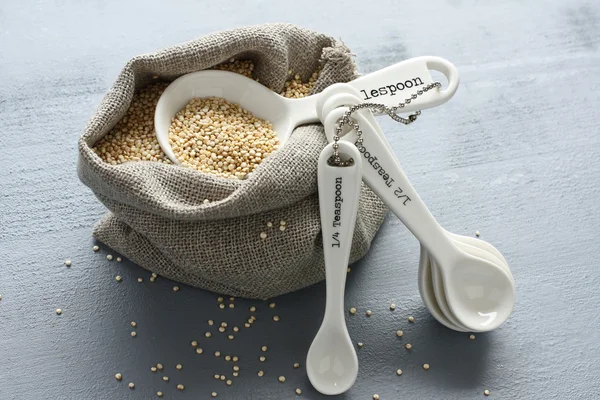 Grão de quinoa em pequeno saco de serapilheira e colheres de medição de porcelana — Fotografia de Stock