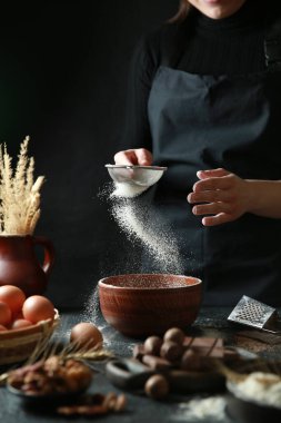 Aşçılık süreci. Un elek yoluyla derin bir seramik kaseye dökülür. Ellerinle önlük pişir. Çikolata, fındık, siyah masadaki yumurtalar. Arkaplan resmi, alanı kopyala