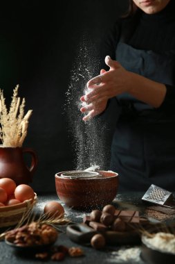 Aşçılık süreci. Un elek yoluyla derin bir seramik kaseye dökülür. Ellerinle önlük pişir. Çikolata, fındık, siyah masadaki yumurtalar. Arkaplan resmi, alanı kopyala