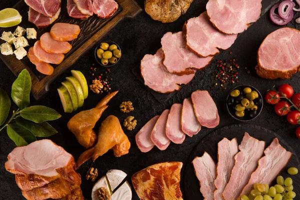 Sausages, bacon, chicken and meat delicacies. Sliced meat, ham with olives, grapes, cheese, cherry tomatoes, herbs, spices on a black background. Background image, copy space, top view, flatlay