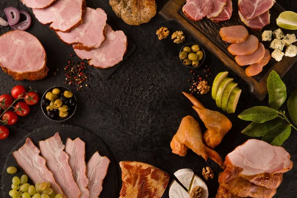 Sausages, bacon, chicken and meat delicacies. Sliced meat, ham with olives, grapes, cheese, cherry tomatoes, herbs, spices on a black background. Background image, copy space, top view, flatlay