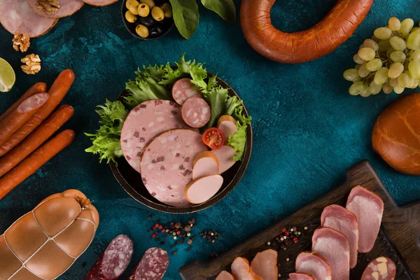Sausages, bacon and meat delicacies. Sliced meat, ham with olives, grapes, lime, cherry tomatoes, herbs, spices and nuts on a dark turquoise background. Background image, copy space, top view, flatlay