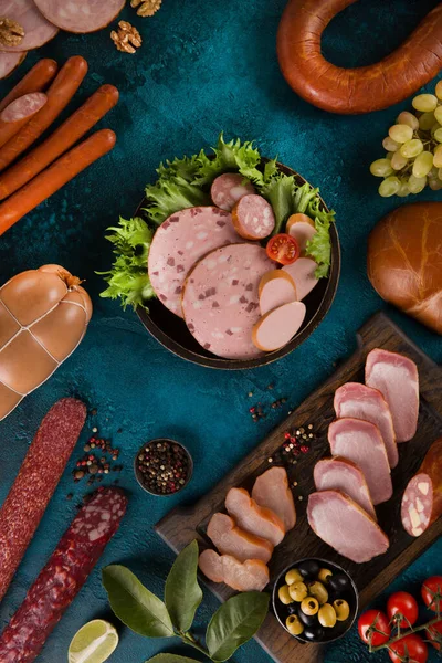 Sausages, bacon and meat delicacies. Sliced meat, ham with olives, grapes, lime, cherry tomatoes, herbs, spices and nuts on a dark turquoise background. Background image, copy space, top view, flatlay