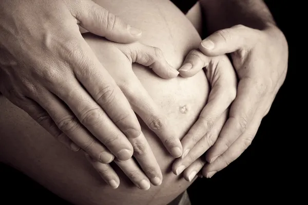 Руки беременной женщины и ее мужа в форме сердца на ней быть — стоковое фото
