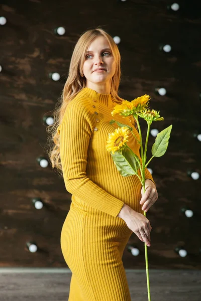a pregnant woman in a yellow dress stands and holds a sunflower