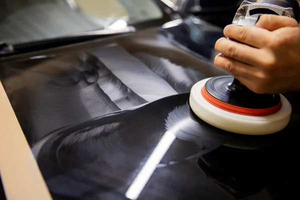 The concept of detailing and polishing cars. The hands of a professional car service worker, with an orbital polisher, polish the black luxury hood of a car in an auto repair shop. car polishing