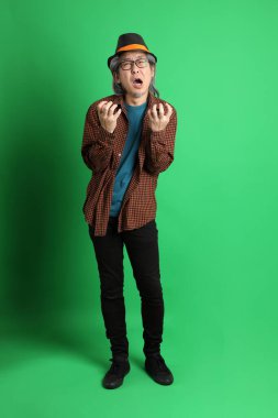 Son sınıf Asyalı adam Şükran Günü renk temalı yeşil arka planda duran turuncu ekose gömlek ve siyah şapka giyiyor..