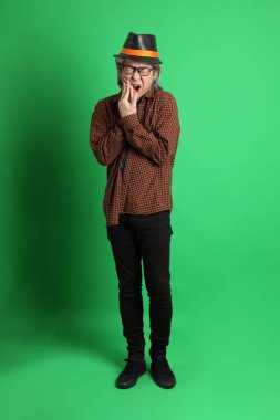 Son sınıf Asyalı adam Şükran Günü renk temalı yeşil arka planda duran turuncu ekose gömlek ve siyah şapka giyiyor..