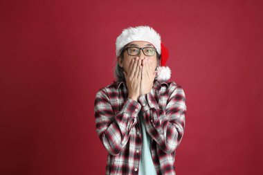 Kıdemli Asyalı adam kırmızı arka planda duran Noel Baba şapkalı kırmızı ekose gömlek giyiyor..