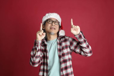 Kıdemli Asyalı adam kırmızı arka planda duran Noel Baba şapkalı kırmızı ekose gömlek giyiyor..