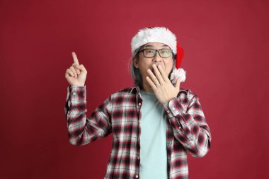 Kıdemli Asyalı adam kırmızı arka planda duran Noel Baba şapkalı kırmızı ekose gömlek giyiyor..
