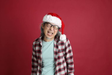 Kıdemli Asyalı adam kırmızı arka planda duran Noel Baba şapkalı kırmızı ekose gömlek giyiyor..