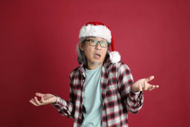 Kıdemli Asyalı adam kırmızı arka planda duran Noel Baba şapkalı kırmızı ekose gömlek giyiyor..