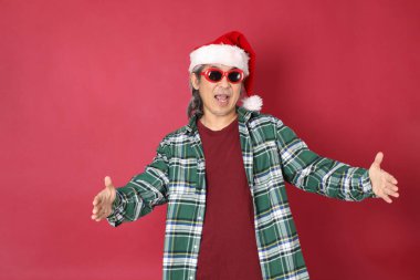 Kıdemli Asyalı adam yeşil ekose gömlek giyiyor ve kırmızı arka planda Noel Baba şapkası var..