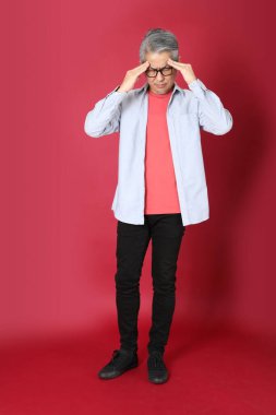 Kırmızı arka planda duran mavi tişörtlü, uzun saçlı kıdemli Asyalı adam..