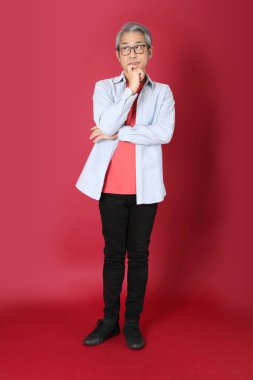 Kırmızı arka planda duran mavi tişörtlü, uzun saçlı kıdemli Asyalı adam..