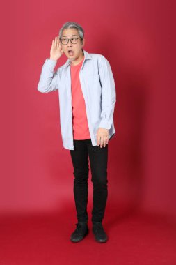 Kırmızı arka planda duran mavi tişörtlü, uzun saçlı kıdemli Asyalı adam..