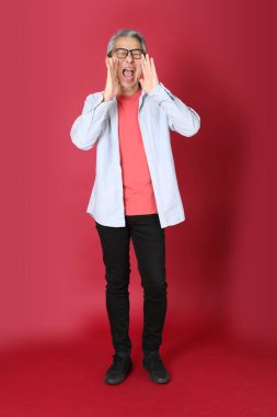 Kırmızı arka planda duran mavi tişörtlü, uzun saçlı kıdemli Asyalı adam..