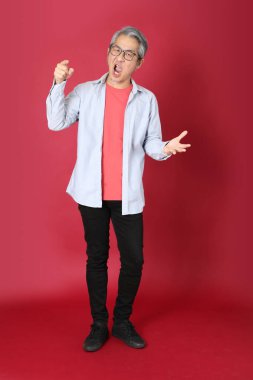 Kırmızı arka planda duran mavi tişörtlü, uzun saçlı kıdemli Asyalı adam..