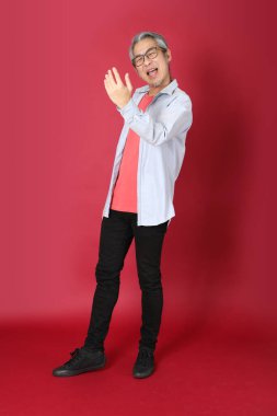 Kırmızı arka planda duran mavi tişörtlü, uzun saçlı kıdemli Asyalı adam..