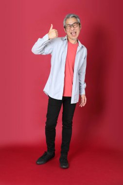 Kırmızı arka planda duran mavi tişörtlü, uzun saçlı kıdemli Asyalı adam..