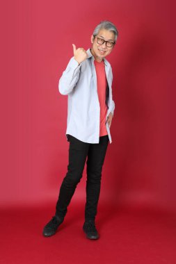 Kırmızı arka planda duran mavi tişörtlü, uzun saçlı kıdemli Asyalı adam..