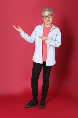 Kırmızı arka planda duran mavi tişörtlü, uzun saçlı kıdemli Asyalı adam..