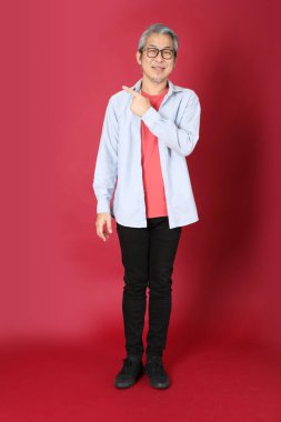 Kırmızı arka planda duran mavi tişörtlü, uzun saçlı kıdemli Asyalı adam..