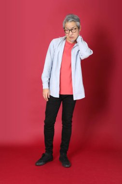 Kırmızı arka planda duran mavi tişörtlü, uzun saçlı kıdemli Asyalı adam..