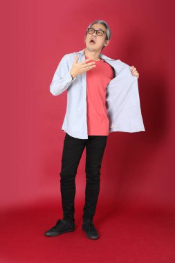 Kırmızı arka planda duran mavi tişörtlü, uzun saçlı kıdemli Asyalı adam..