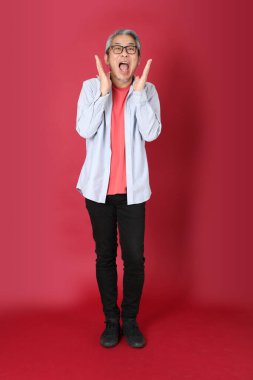 Kırmızı arka planda duran mavi tişörtlü, uzun saçlı kıdemli Asyalı adam..