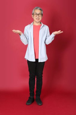 Kırmızı arka planda duran mavi tişörtlü, uzun saçlı kıdemli Asyalı adam..