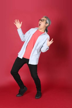 Kırmızı arka planda duran mavi tişörtlü, uzun saçlı kıdemli Asyalı adam..