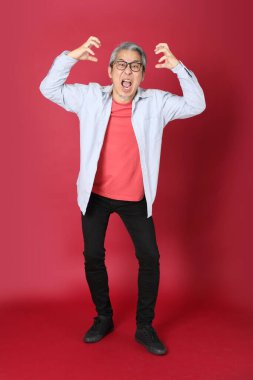 Kırmızı arka planda duran mavi tişörtlü, uzun saçlı kıdemli Asyalı adam..