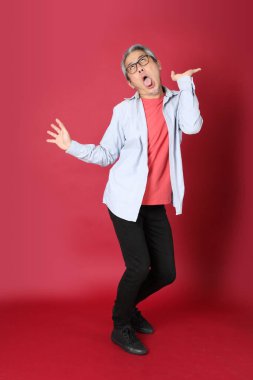 Kırmızı arka planda duran mavi tişörtlü, uzun saçlı kıdemli Asyalı adam..