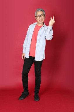 Kırmızı arka planda duran mavi tişörtlü, uzun saçlı kıdemli Asyalı adam..