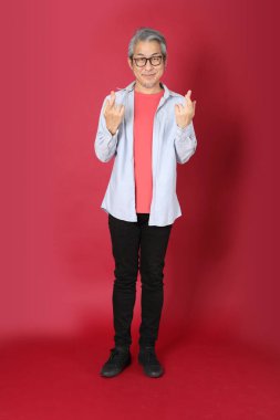 Kırmızı arka planda duran mavi tişörtlü, uzun saçlı kıdemli Asyalı adam..