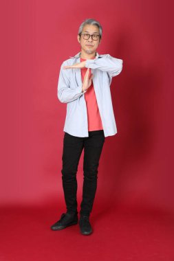 Kırmızı arka planda duran mavi tişörtlü, uzun saçlı kıdemli Asyalı adam..