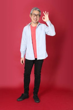 Kırmızı arka planda duran mavi tişörtlü, uzun saçlı kıdemli Asyalı adam..