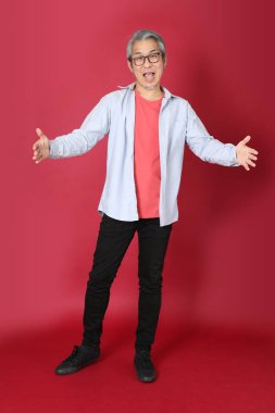 Kırmızı arka planda duran mavi tişörtlü, uzun saçlı kıdemli Asyalı adam..